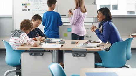 photo of teacher sitting at desk with two students. desk is modern and modular with blue chairs and white boards attached to the side of the desk