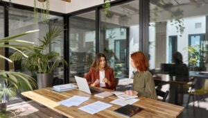 employee in an open floor plan office meeting with a client wooden desk, green plants in the corners, natural lighting