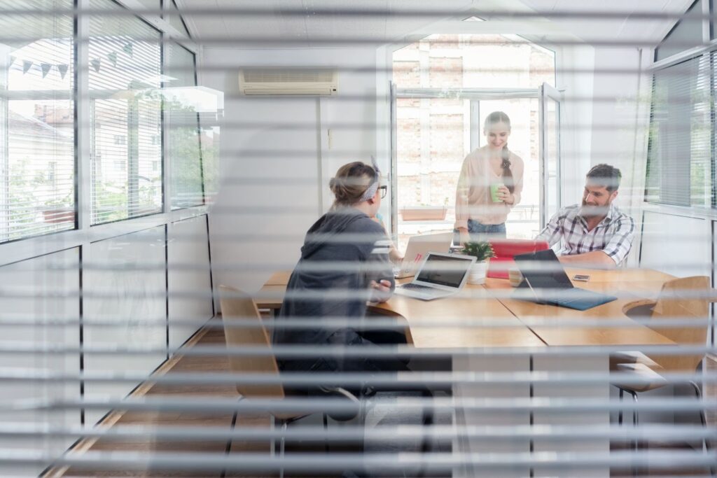 view through open blinds of conference room where 3 coworkers are talking. office window treatments on windows. 